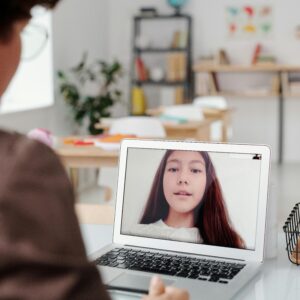 Teacher using laptop for virtual lesson with female student on screen in classroom setting.